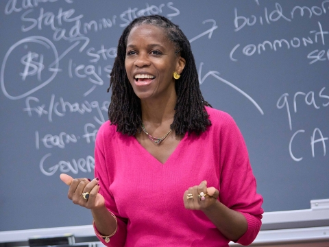 Olatunde Johnson in bright pink dress standing before blackboard covered in writing