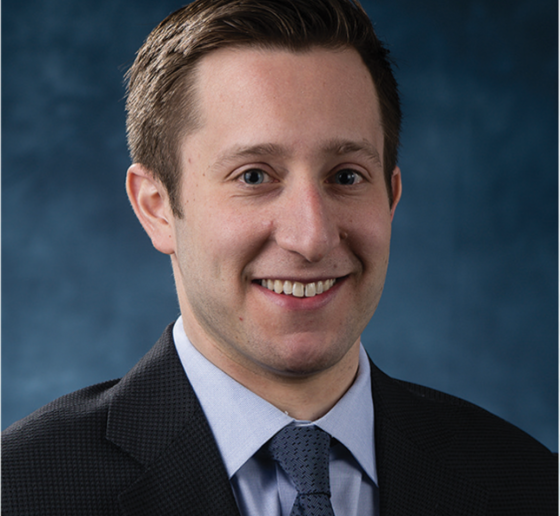 Head shot of man in dark jacket and blue tie.
