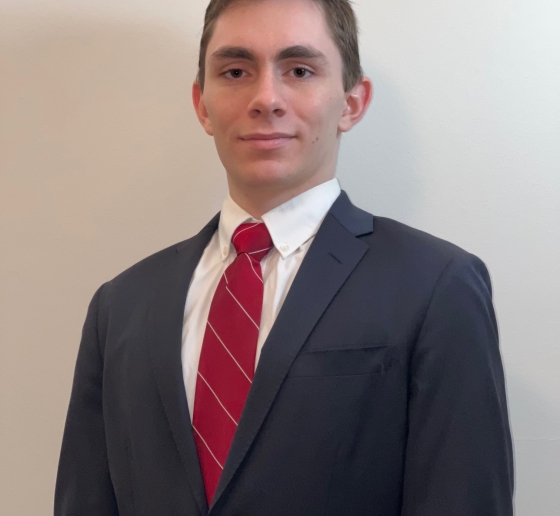 Student in gray suit and red tie stands in front of a white wall