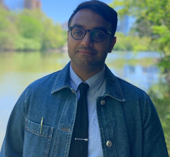 Man wearing glasses, a denim jacket, white shirt, and dark tie