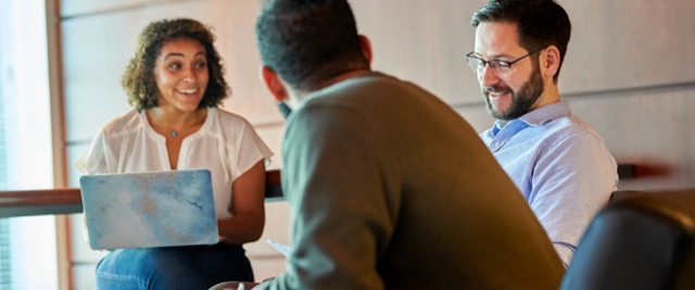 Three students speak around laptops in a lounge area.