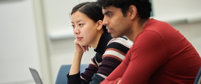 Two students sit in a lecture hall with their laptops open.