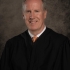 Pictured above is Richard J. Sullivan in traditional judge attire. He is wearing a white button up and copper tie. He has fair skin and grey hair. He is smiling wide. 