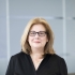 A white woman with blonde hair wears a black shirt and black-framed glasses.