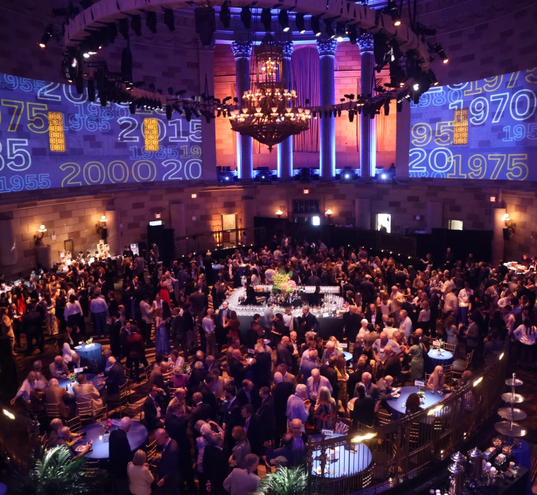 Wide shot of room filled with people at a cocktail party with Columbia logo projections on the walls. 