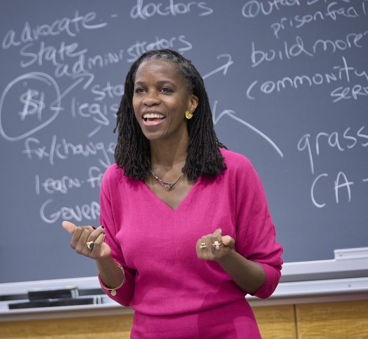Olatunde Johnson in bright pink dress standing before blackboard covered in writing