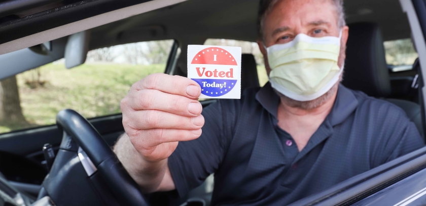 Man wearing a surgical mask for COVID-19 pandemic holds a sticker that says "I VOTED"