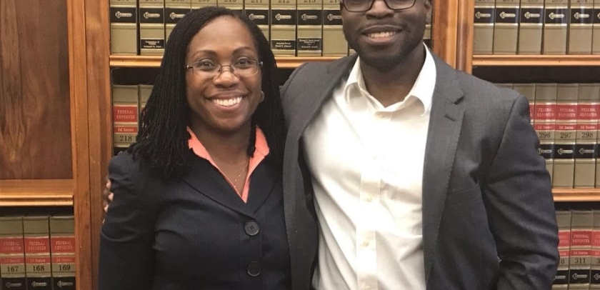 A woman and man both in dark suits, in front of law books 