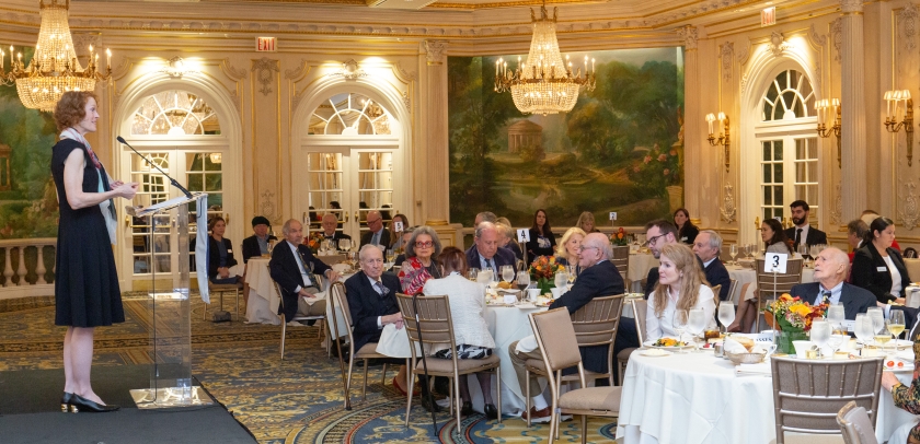 Person on a stage speaking to audience members seated at tables in a dining room