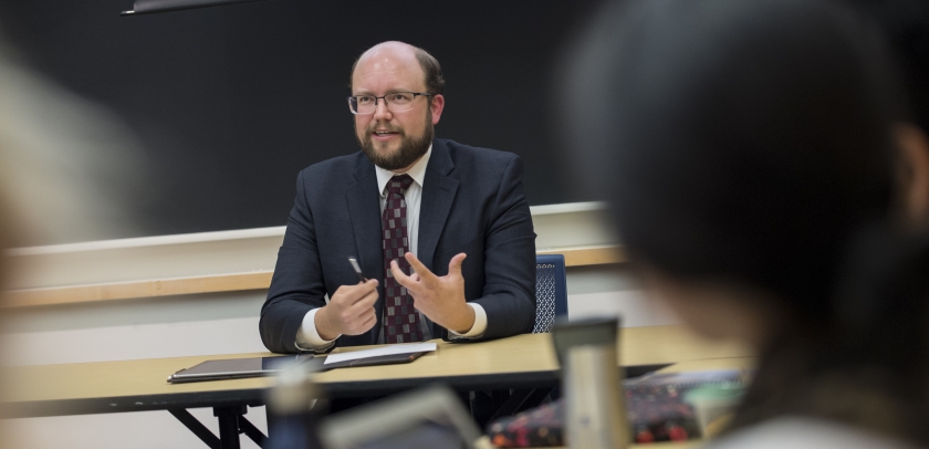 Man in glasses and suit at table in front of a classroom