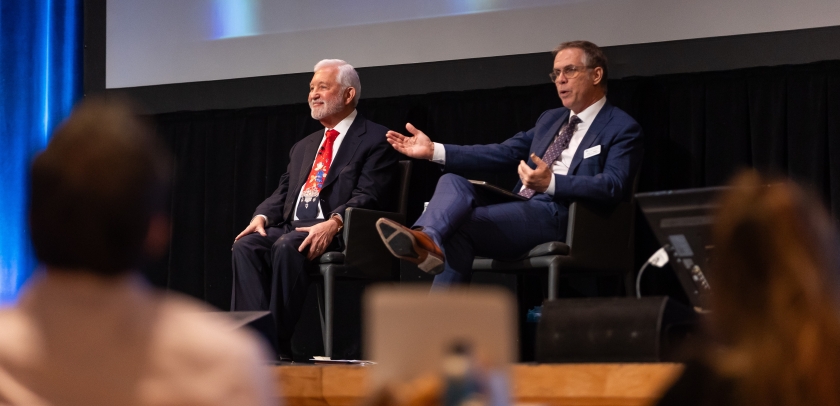 Two men seated onstage for panel discussion, both in suits. 