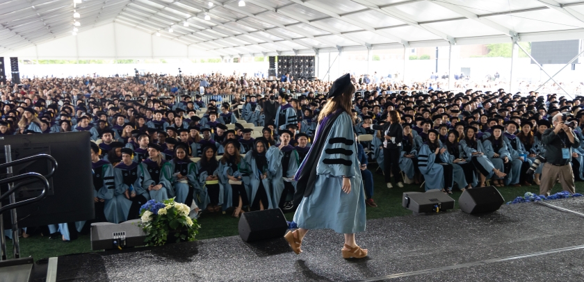 Woman in academic regalia crosses stage before large audience under enormous tent