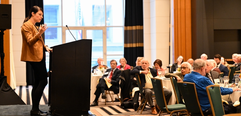 Woman standing at podium speaking to audience seated at tables
