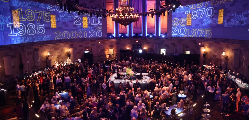 Wide shot of room filled with people at a cocktail party with Columbia logo projections on the walls. 