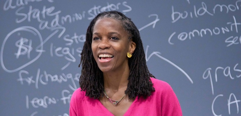 Olatunde Johnson in bright pink dress standing before blackboard covered in writing