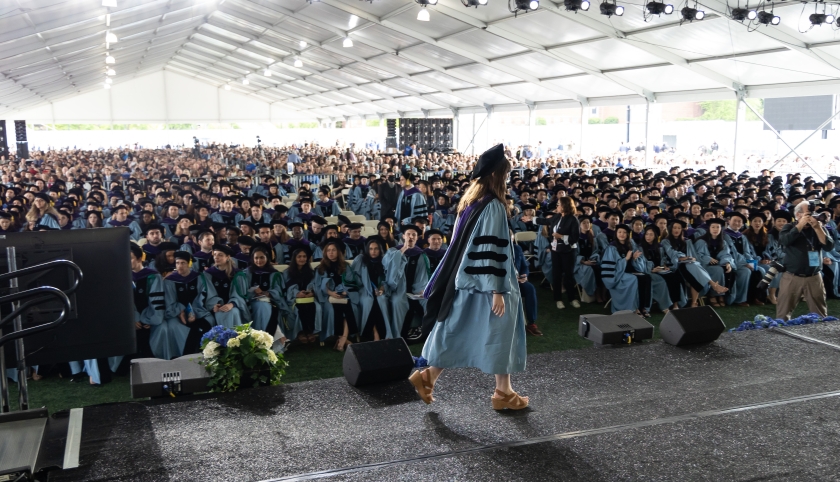 Woman in academic regalia crosses stage before large audience under enormous tent