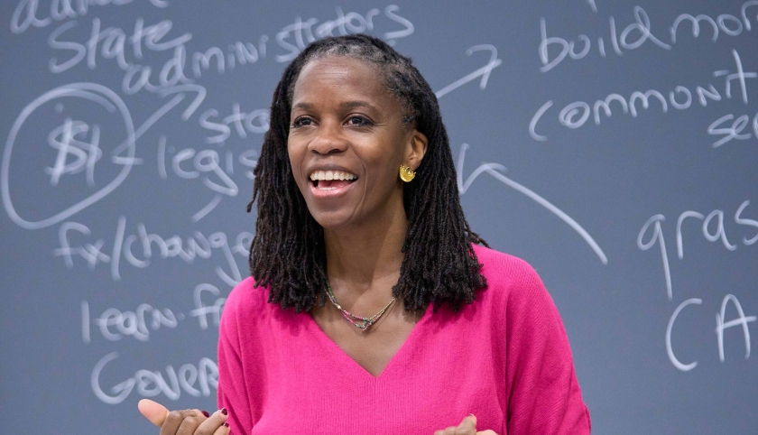 Olatunde Johnson in bright pink dress standing before blackboard covered in writing
