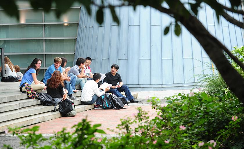 Framed by trees, a group of students sit on the stairs of Revson Plaza.