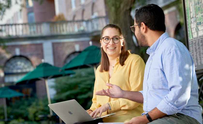 Two smiling students sit on a bench in the Wien courtyard with a laptop.