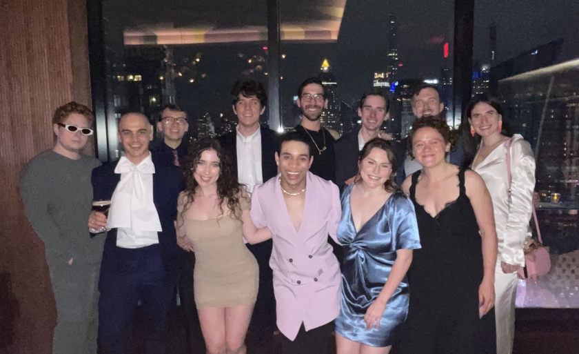 A group of students in cocktail attire stand in front of a city skyline at night.