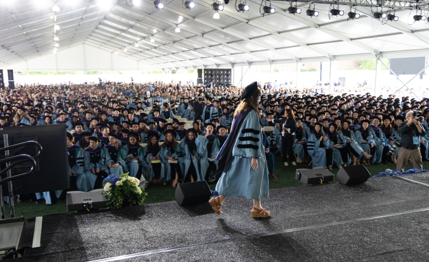 Woman in academic regalia crosses stage before large audience under enormous tent