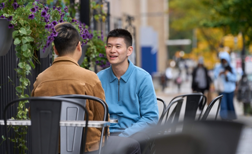 Two students sit at outdoor cafe tables near campus.