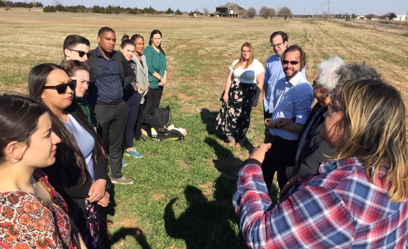 Students stand in a field of grass while a woman speaks to them.