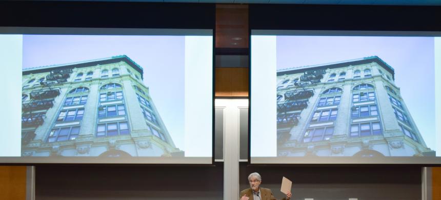 Professor Vincent Blasi gestures in front of a screen featuring a multistory building.
