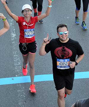 Elora Mukherjee crossing a blue finish line, smiling and raising her hands