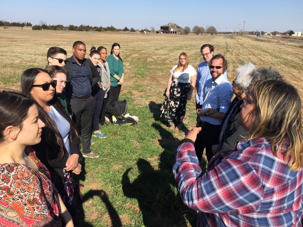 Students stand in a field of grass while a woman speaks to them.