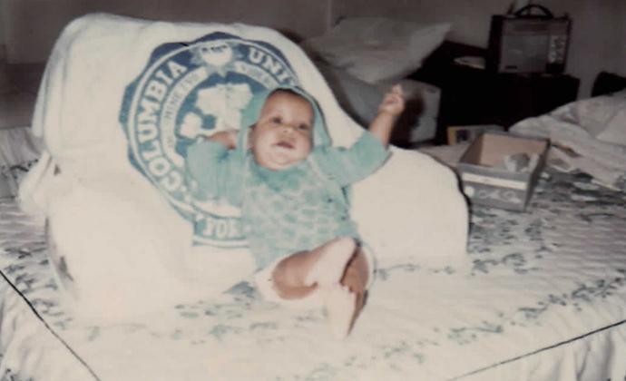 A baby wearing a blue sweater reclines on a pillow with the Columbia logo on it.