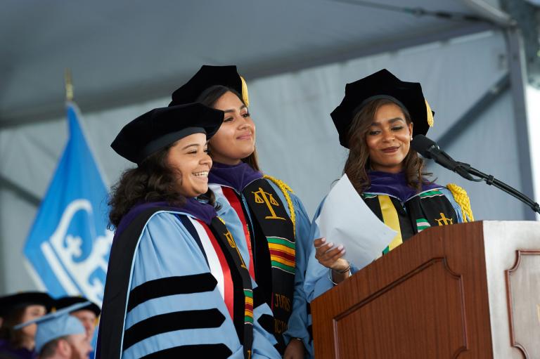 Three graduates in blue caps and gown speak at a podium.