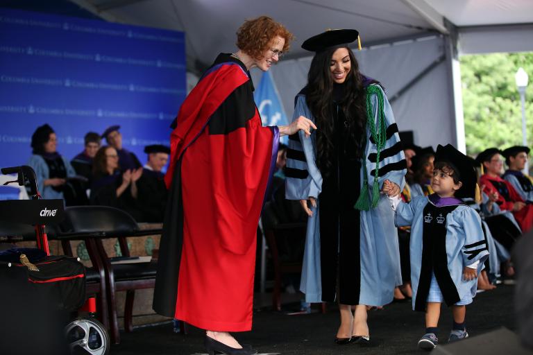 A student in cap and gown holds the hand of a young child in matching regalia on the stage.