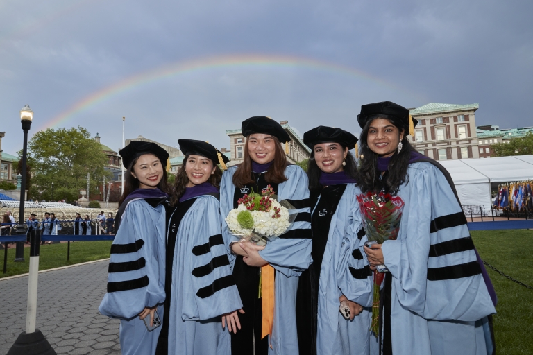 Five students in caps and gowns with rainbow in background