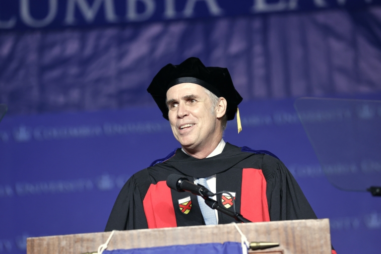 Professor in black cap and gown with red sash at podium