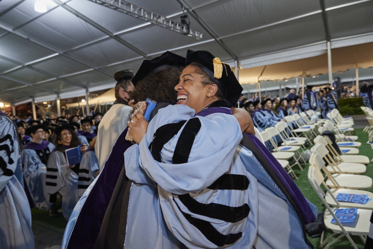 Two woman in caps and gowns hugging at graduation under tent