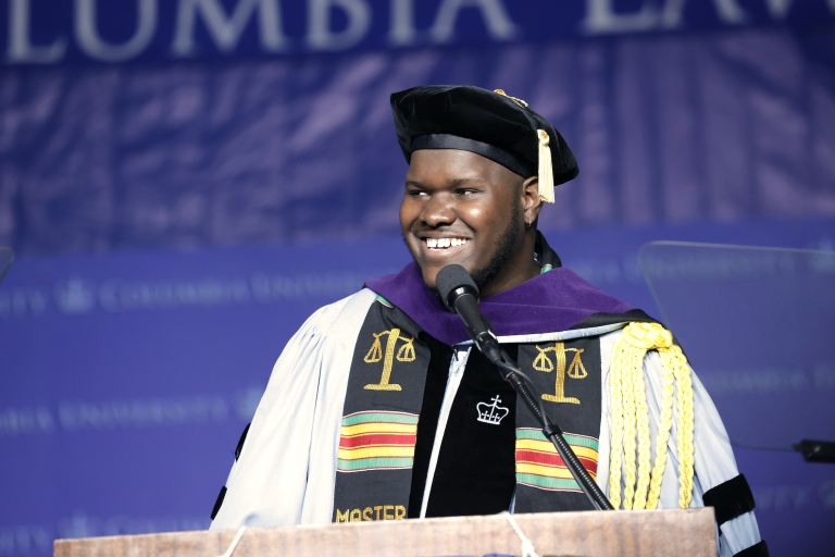 Man in cap and gown with colorful sash at podium