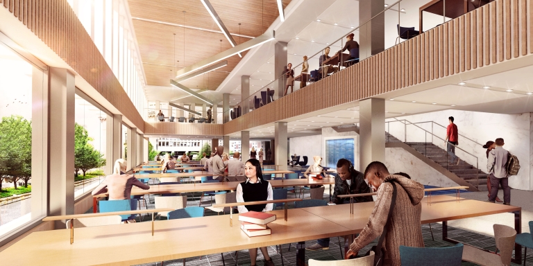 Students sit at long tables next to a bank of windows and beneath a mezzanine.