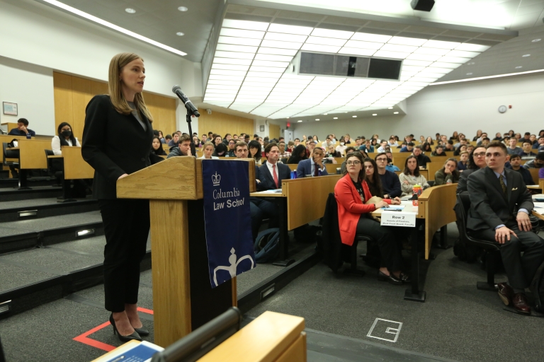 Audience members look at a woman standing at a podium.