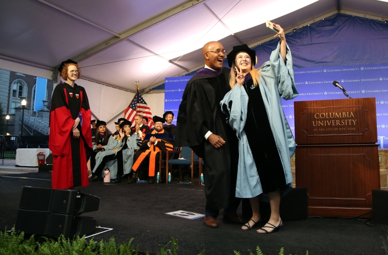 Woman in academic gown takes selfie onstage with man in academic gown