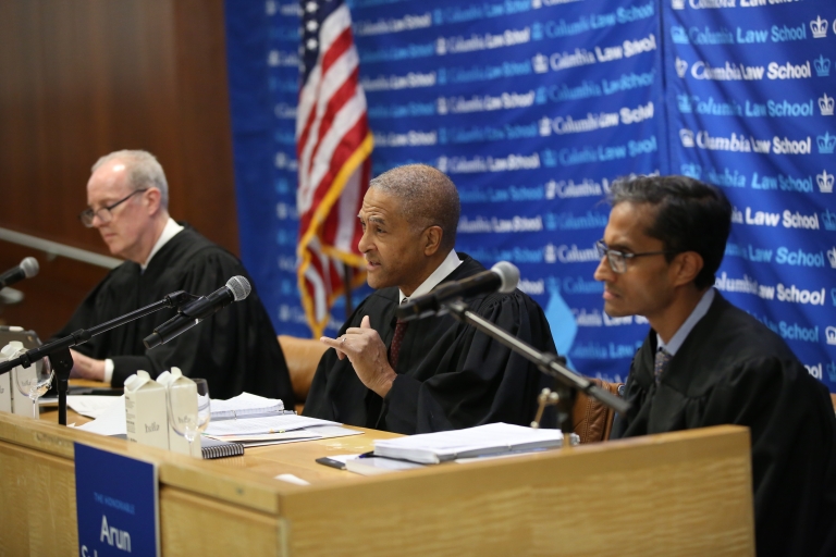 Three men in judges' robes seated behind a desk and speaking into microphones 