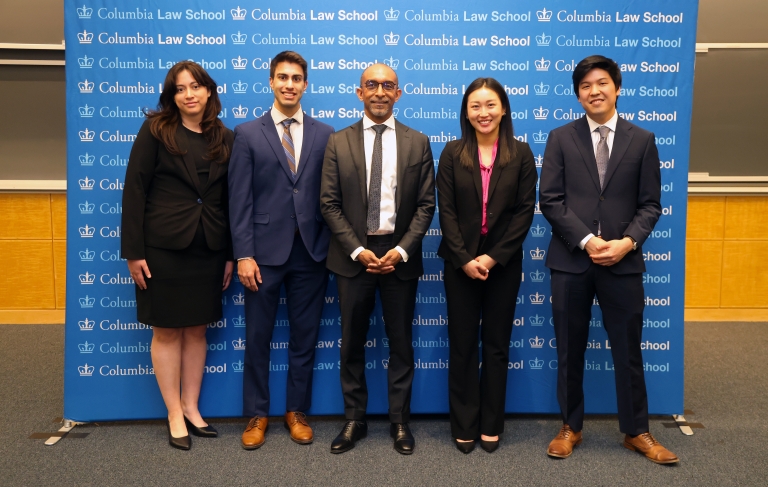 Five people stand in front of a sign with the Columbia Law School logo repeated
