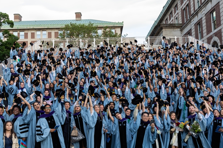 Photo of cheering students in the bleachers at the Law School's graduation ceremony. They are wearing blue gowns and black graduation caps, and some are tossing their caps into the air.