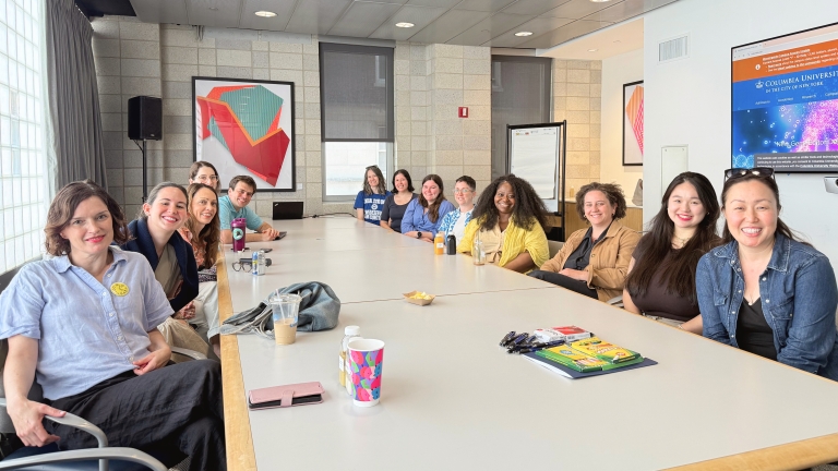 The PI/PS Office staff sits around a long conference table. Everyone smiles at the camera. There are cups, markers, and other items scattered across the table.