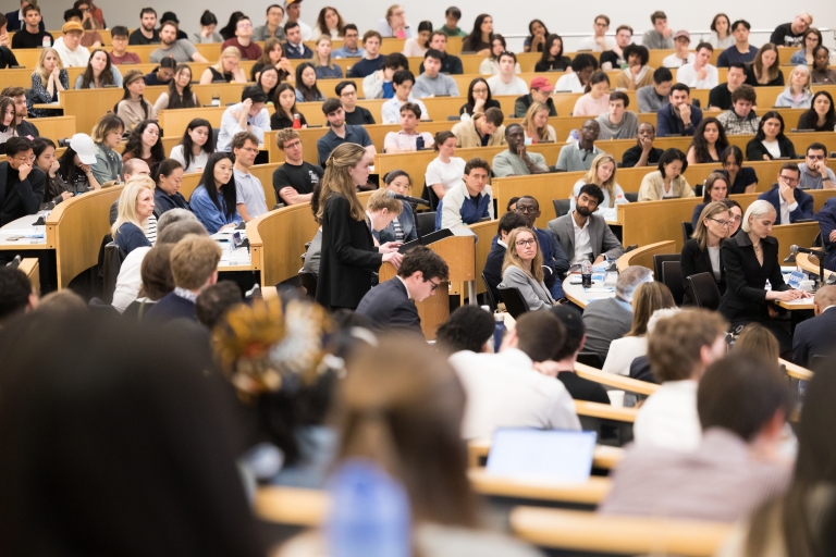 A student stands at a podium in the center of classroom with students seated around her.