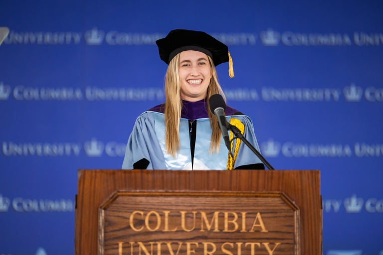 Student Senate president Celeste Woloshyn ’26 in academic regalia stands behind a Columbia University podium