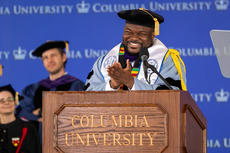 J.D. class speaker, Shaquille Profitt ’26, in academic regalia stands  behind a wood podium with the words Columbia Univeristy on itbehind