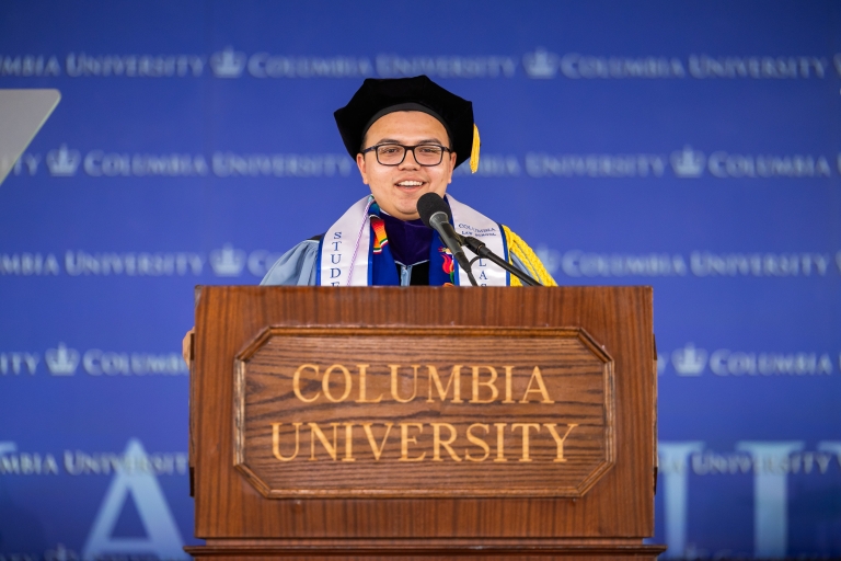 Graduation Committee Co-Chair Amado Aztlán Castillo ’26 in academic regalia standing behind a wood podium with the words Columbia University on it