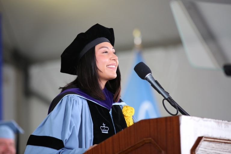Melissa De La Torre ’26 in academic regalia before a microphone