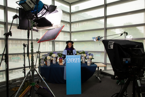 Woman in academic robes behind podium surrounded by camera equipment